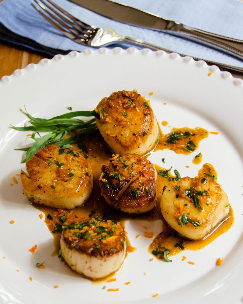 Pan-Seared Scallops with orange tarragon sauce on a white plate with knife and fork in background.