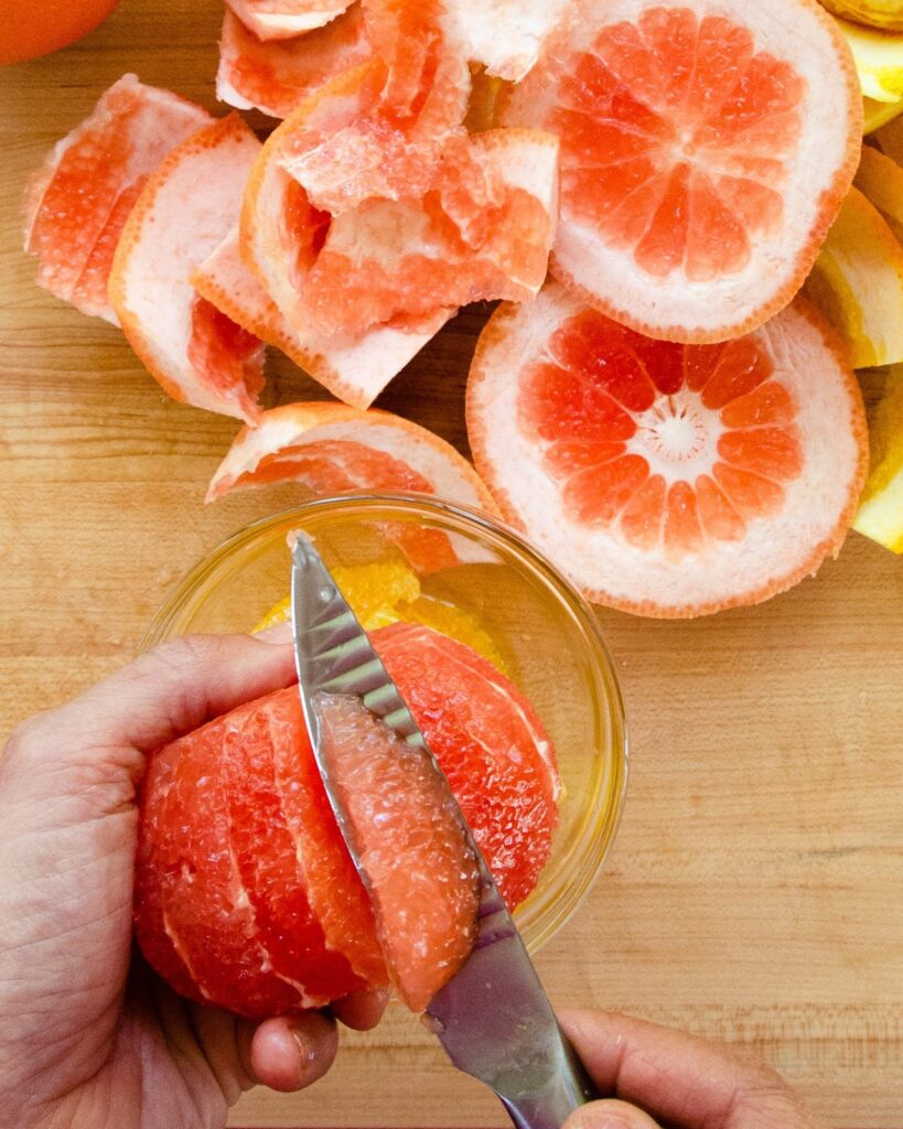A segment of grapefruit on a paring knife with peels in the background.