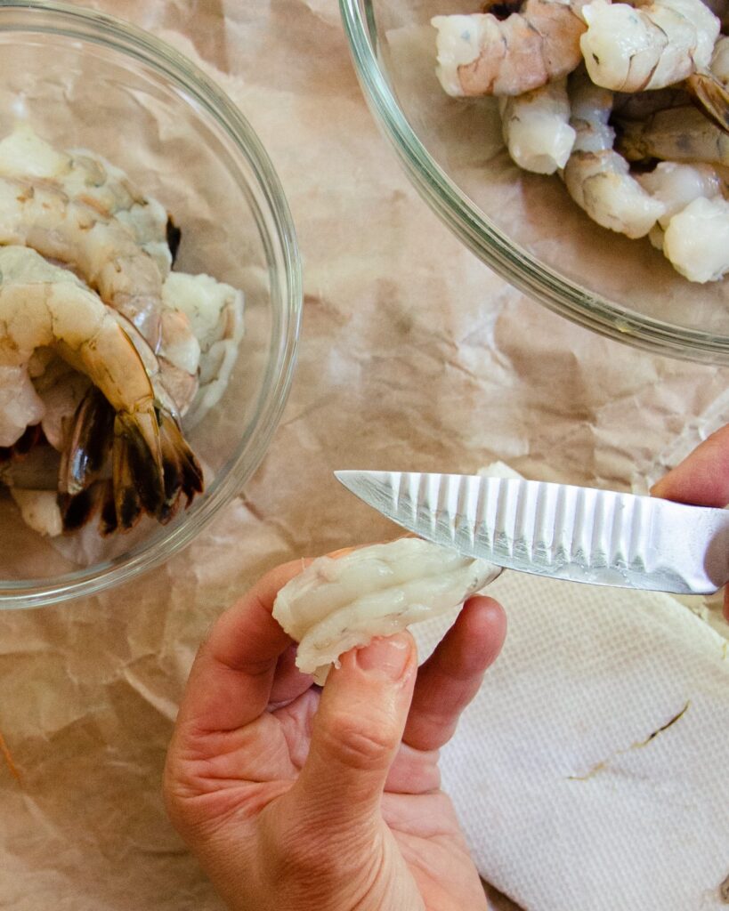 A knife showing a peeled and deveined shrimp with paper bowl and the removed "vein" below.