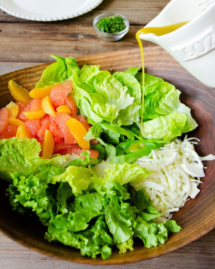 Preparing a grilled shrimp and citrus salad - Vinaigrette being poured on greens, fennel and citrus segments in a large wooden salad bowl.