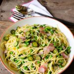 Spaghetti with zucchini, peas, radishes and mint in a serving bowl on a wooden table with parmesan cheese on the side.