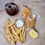 Sheet pan fish and chips on brown paper with malt vinegar and tartar sauce.