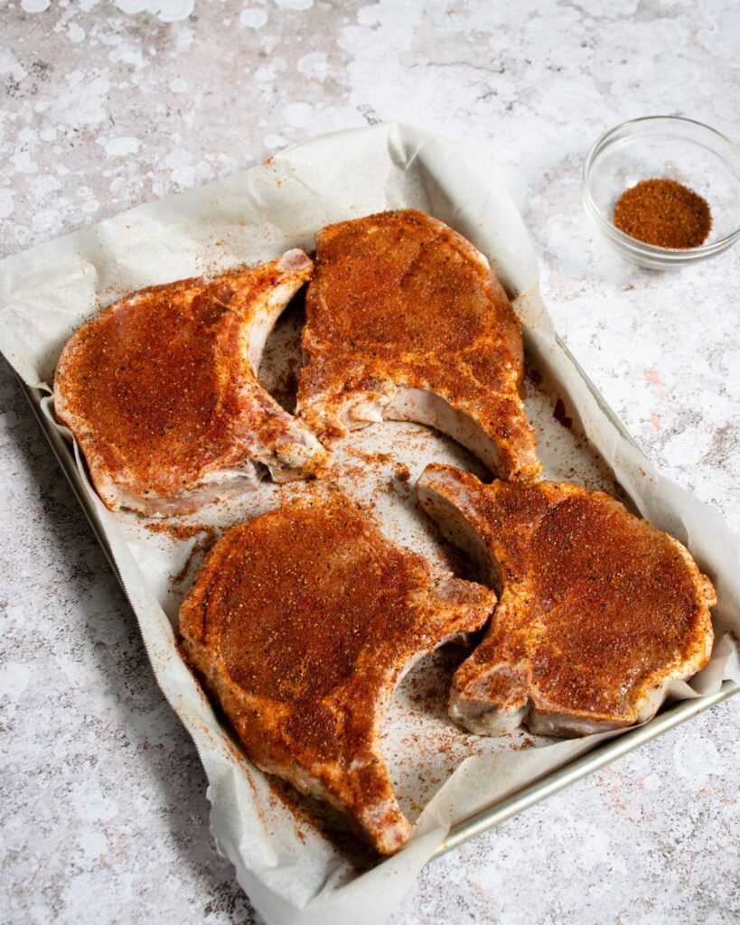 Grilled Brined Pork Chops Pork chops on a parchment-lined quarter sheet pan with a spice rub.