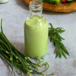 Green Goddess Dressing in a bottle with fresh herbs beside it and a salad in the background.