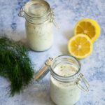 Lemon Dill Buttermilk Dressing in jars with a halved lemon and some fresh dill on a blue marble counter.