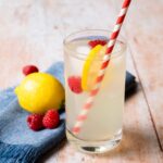 A glass of lemonade on a wooden table with a red and white striped straw, a lemon and some raspberries.