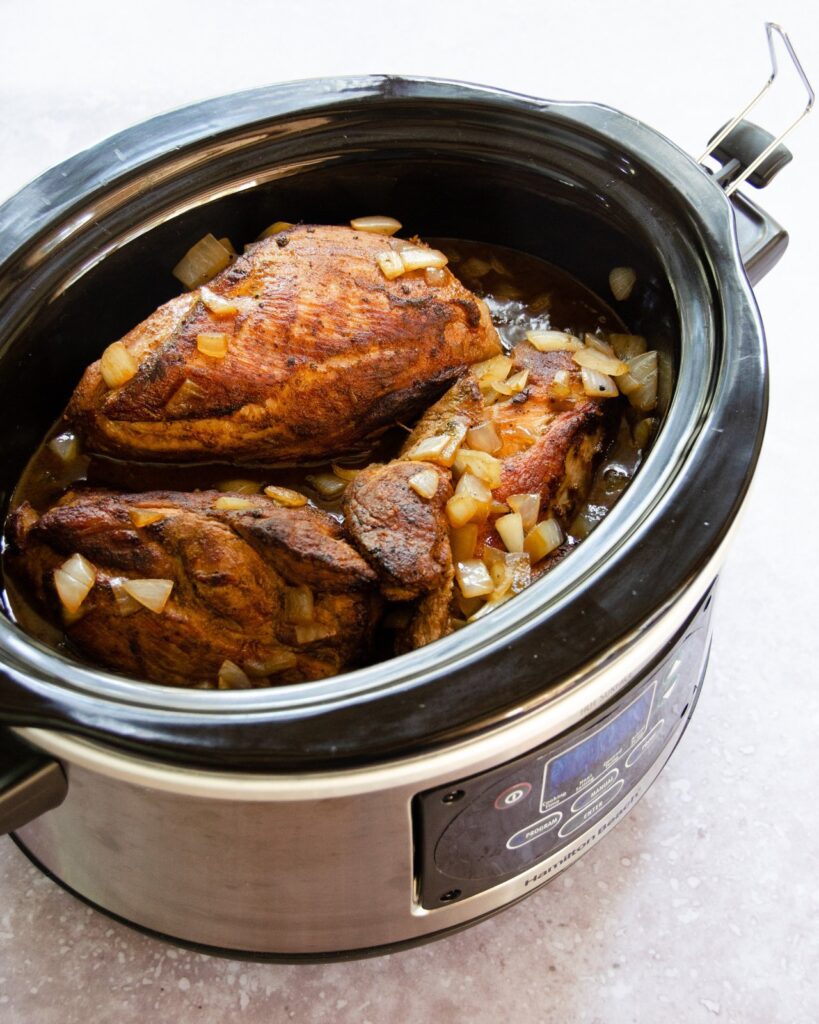 Pieces of browned pork butt in a slow cooker with onions and liquid.