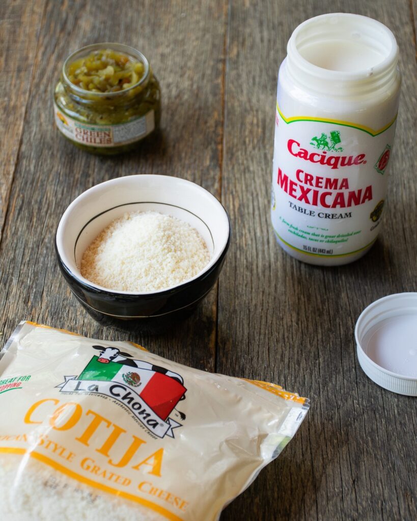 Cotija cheese, mexican creme and canned green chilis in containers on a wooden table.