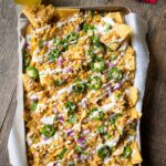 Mexican Street Corn Nachos on a parchment lined baking sheet on a wooden table with a beer and red napkins near by.