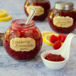 Jar of raspberry limoncello jam on a counter with a spoon, lemons and raspberries scattered around.