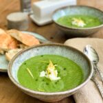 2 bowls of Zucchini Basil Gazpacho on a wooden table with bread and butter in the background.