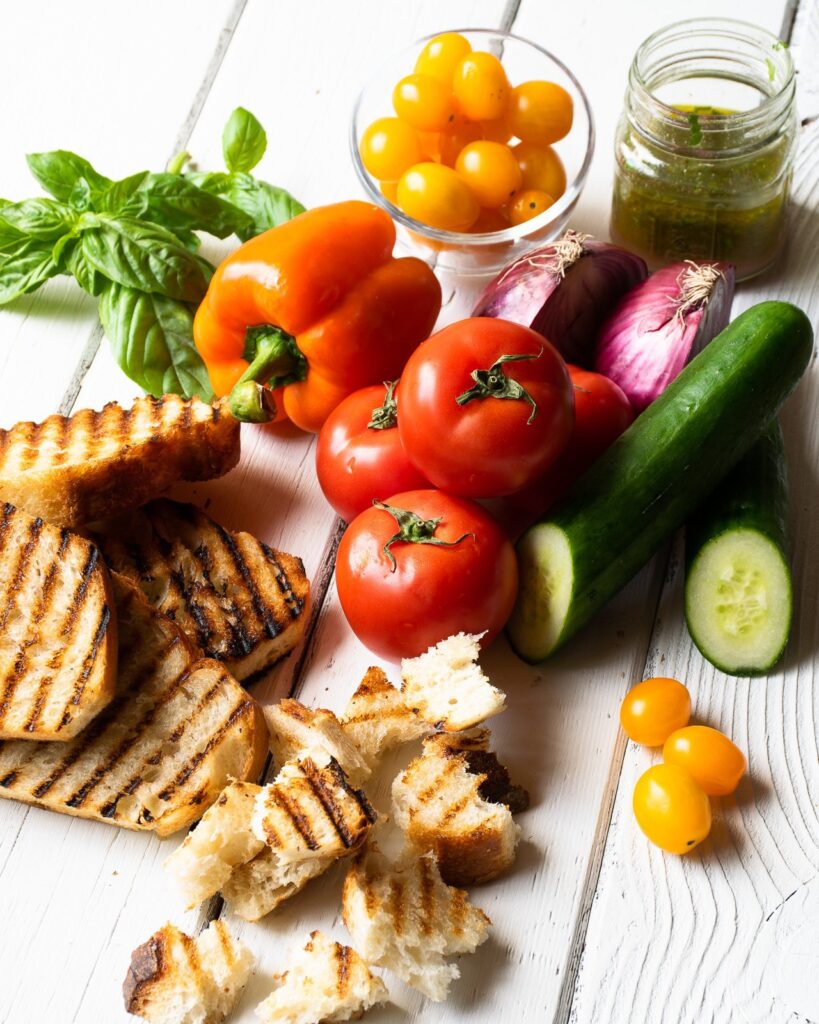 Grilled bread, tomatoes, peppers, cucumber, red onion, basil and dressing on a wooden table.