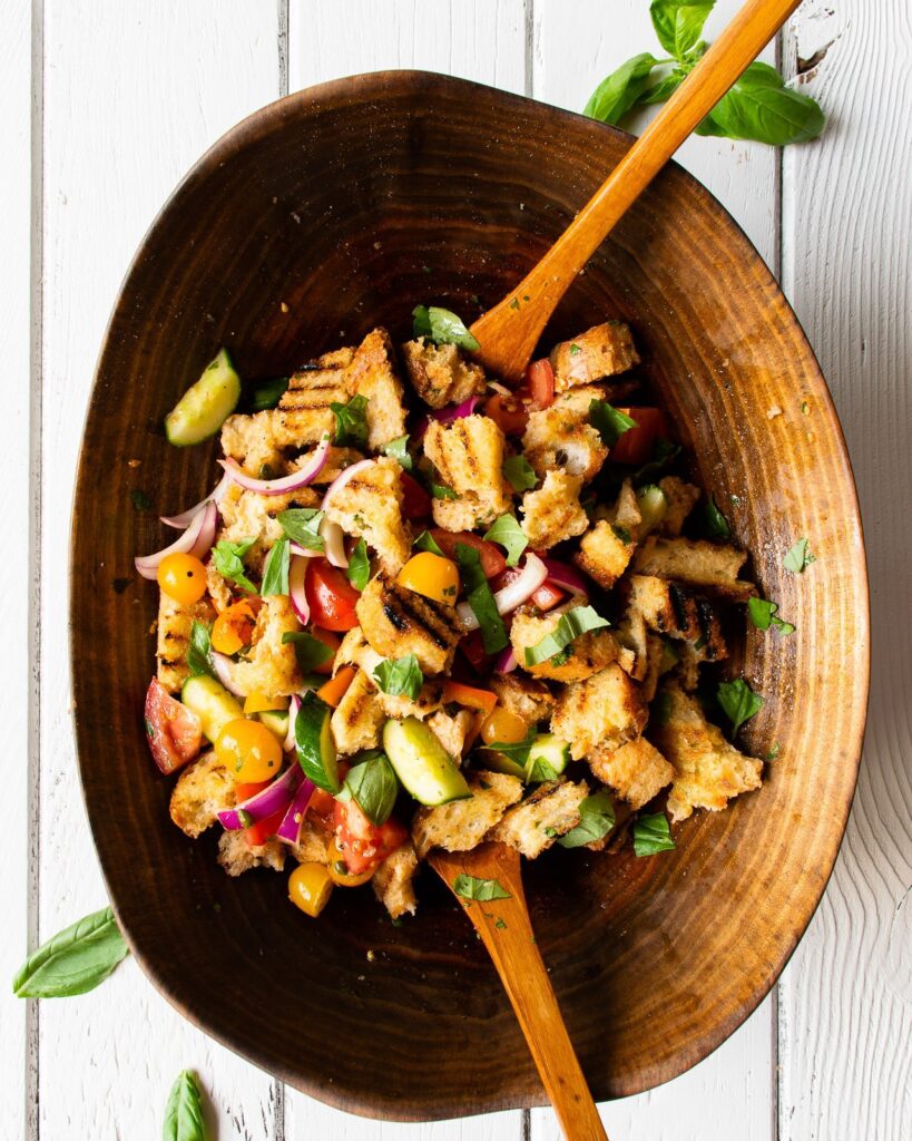 A large wooden bowl with grilled panzanella salad and two serving tools on a white wooden table.