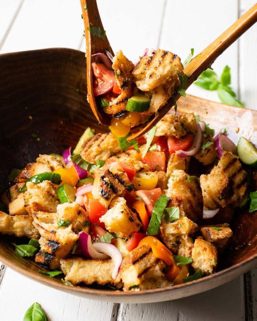 Two salad servers lifting grilled panzanella salad out of the bowl.
