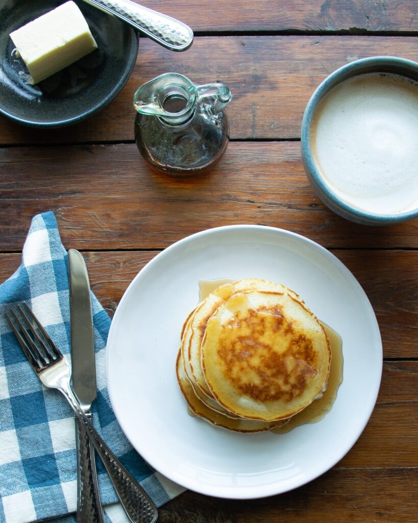 Pancakes on a white plate on a wooden table with coffee, maple syrup and butter.