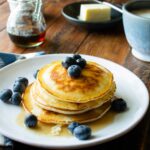 Pancakes with maple syrup and blueberries on a white plate on a wooden table with maple syrup, butter and coffee in the background.