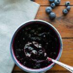 Blueberry Cinnamon Compote in a bowl with a spoon and several blueberries on a wooden table.