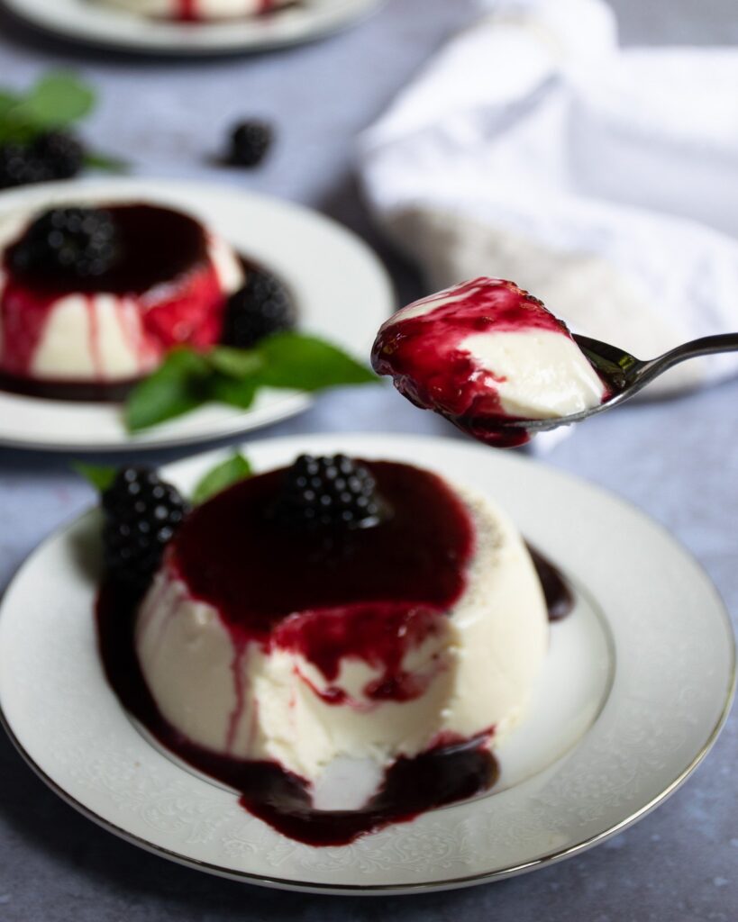 Panna Cotta with Blackberry Brandy Sauce on a plate with a spoon scooping out a mouthful.