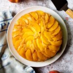Upside Down Peach Cake on a white cake plate with a pie server, some peaches and a blue and tan towel near by.
