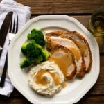 Slices of roast turkey on a white plate with mashed potatoes and broccoli on a wooden table with knife, fork and napkin.