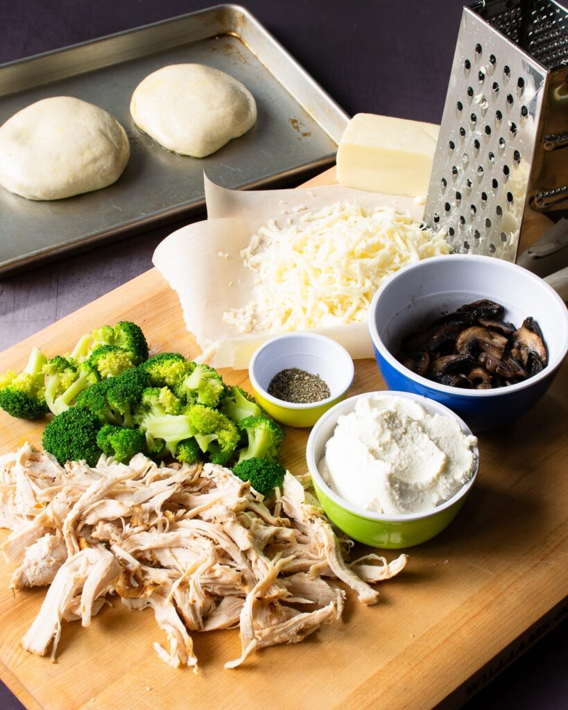 Ingredients on a cutting board - turkey, broccoli, mushrooms,, cheese, spices and pizza dough in the background.