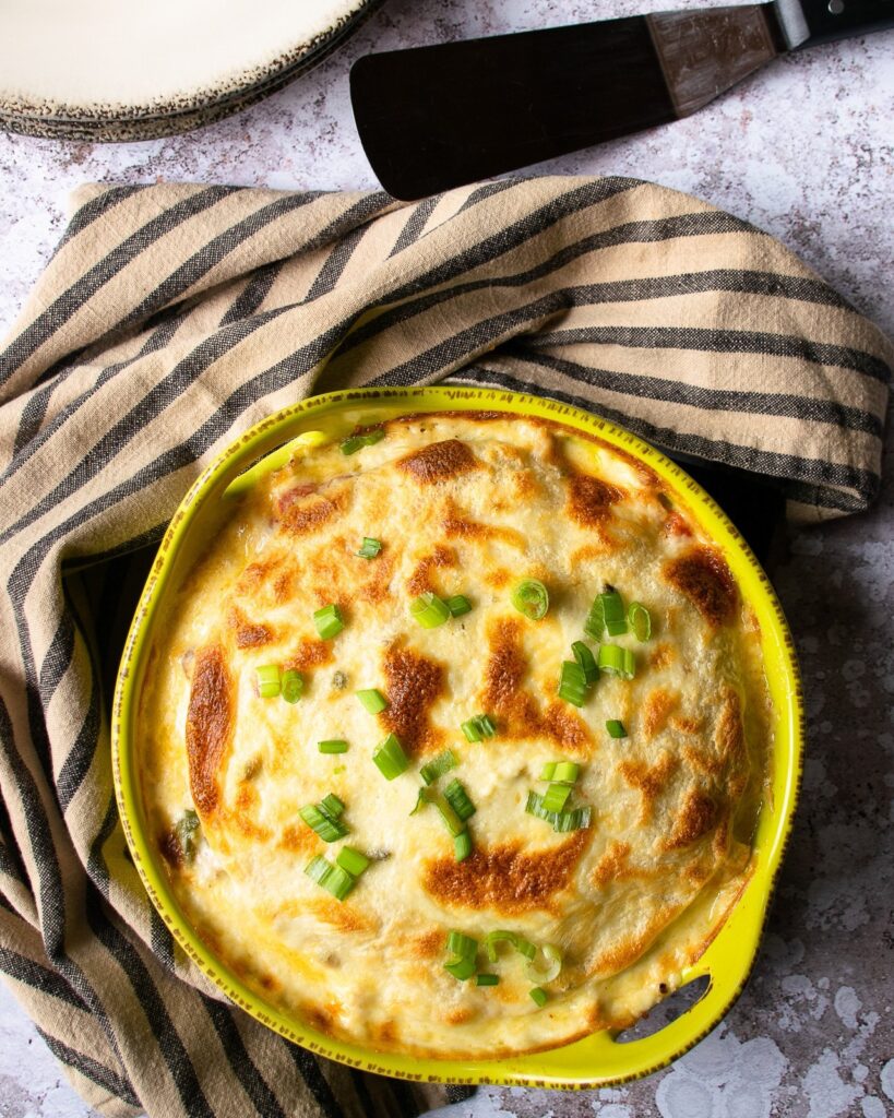 White Turkey Enchilada Pie in a green casserole on a counter with a brown and black towel and server near by.