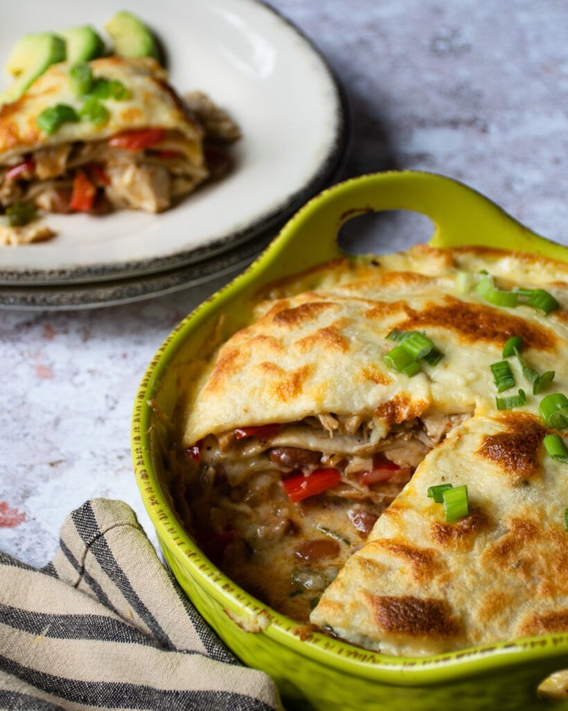 Turkey enchilada pie in a green casserole with a wedge removed and sitting on a white plate in the background.