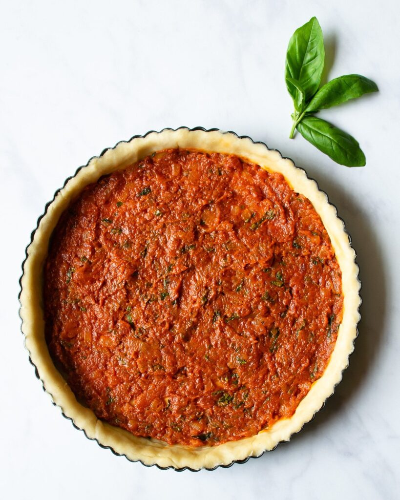Tart shell with tomato sauce in the bottom of it and a random basil leaf near by.