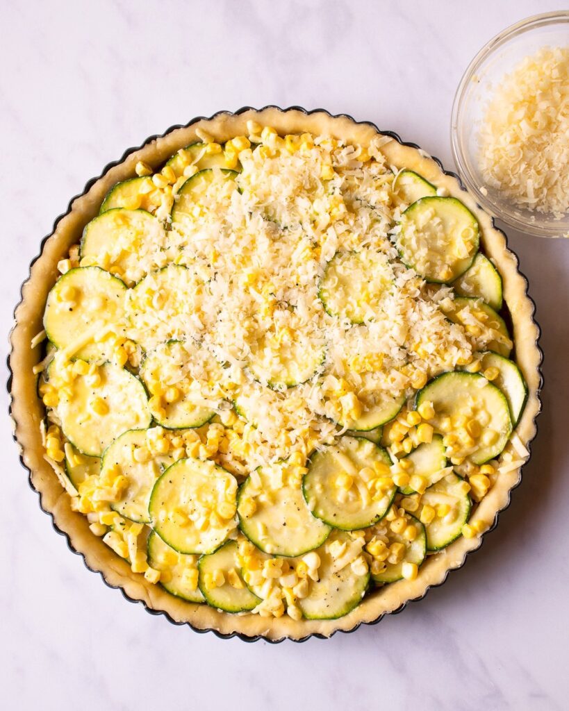 Zucchini, corn and cheese in a tart shell, unbaked with a bowl of cheese nearby.