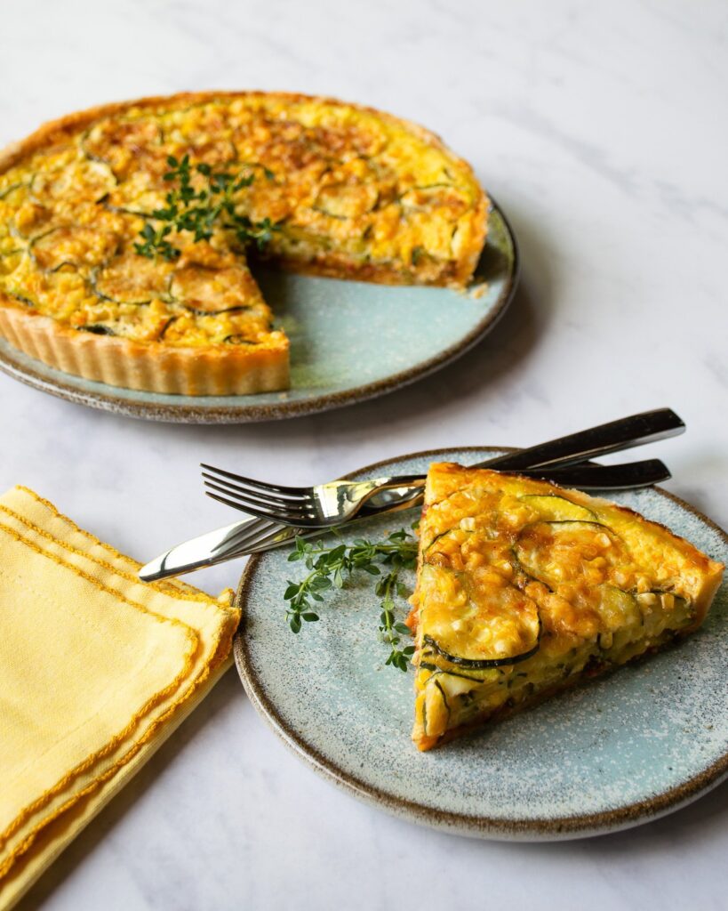 A piece of zucchini and corn tart on a blue plate with a yellow napkin and the rest of the tart in the background.