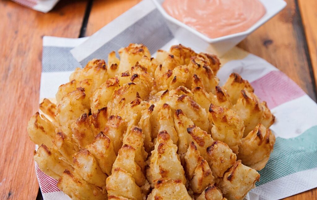 Blooming onion on a colored napkin on a wooden table with a dipping sauce near by.