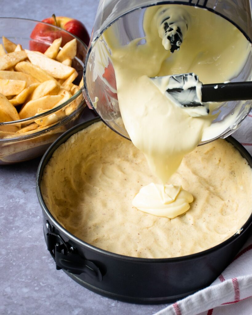 Pouring batter into a dough lined springform pan with sliced apples in the background.