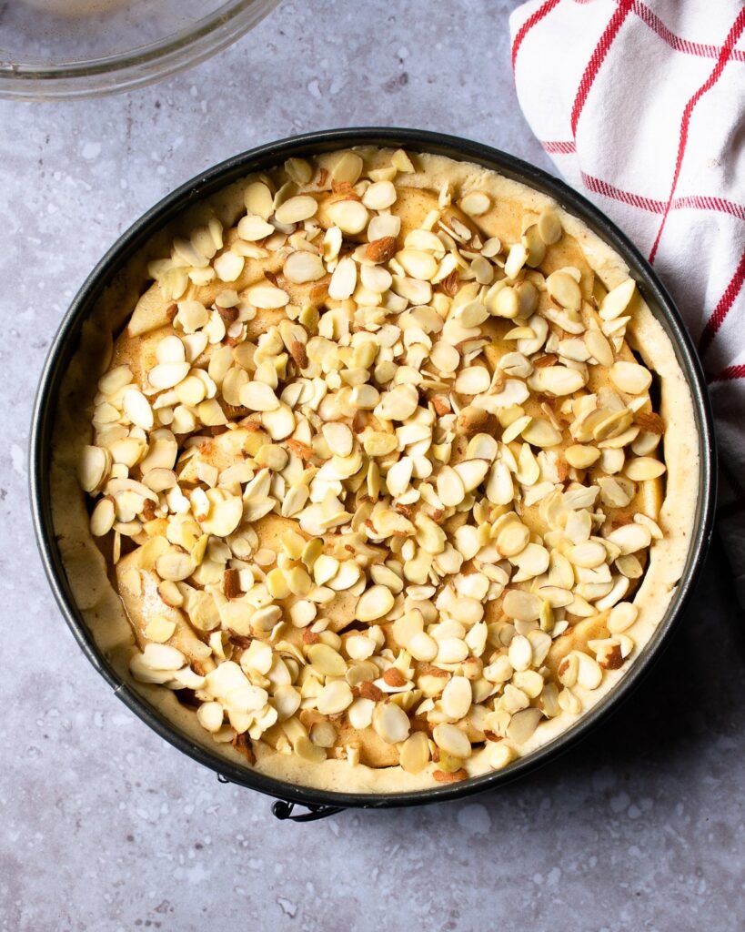 Almonds on top of apples in a springform pan with a red and white windowpane towel near by.