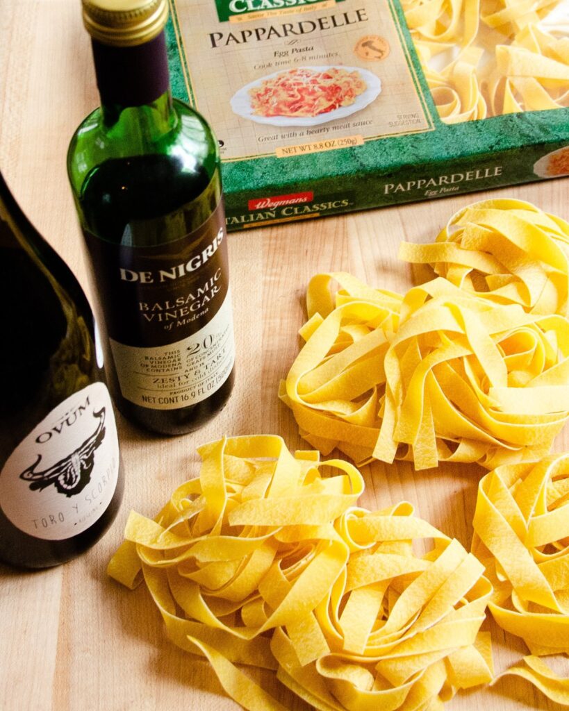Nests of dried pappardelle on a wooden cutting board next to a bottle of white wine and balsamic vinegar.
