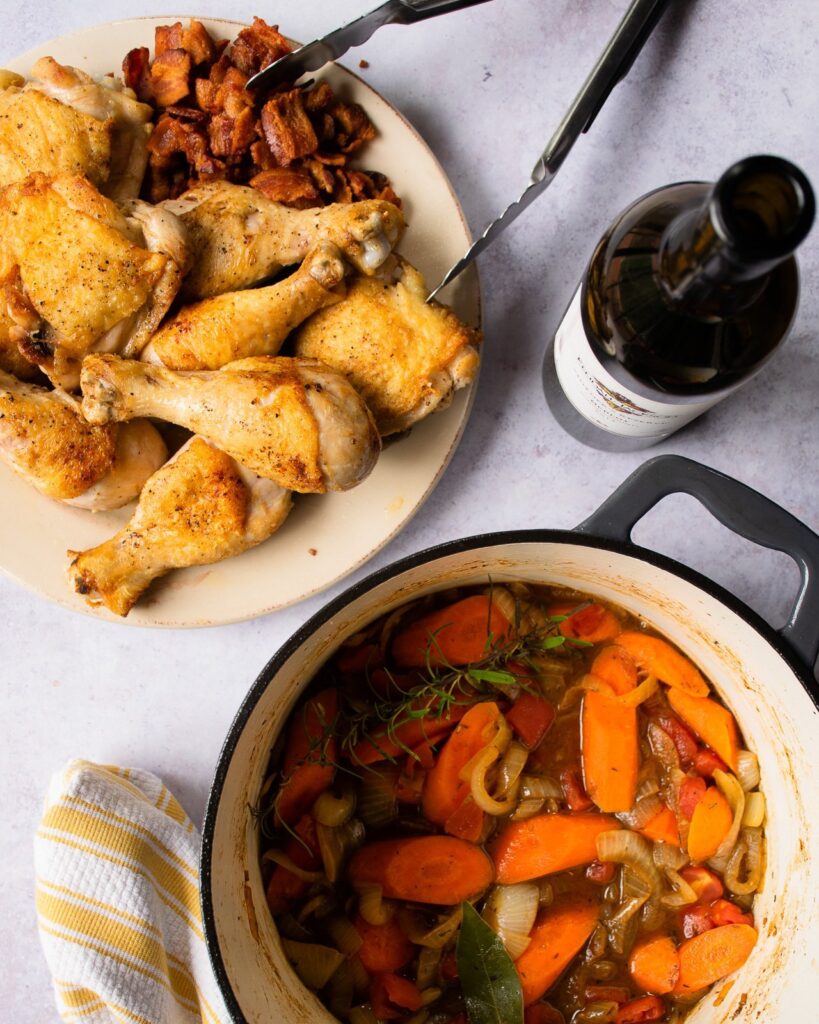Browned chicken on a plate, vegetables in a Dutch oven and a bottle of red wine.