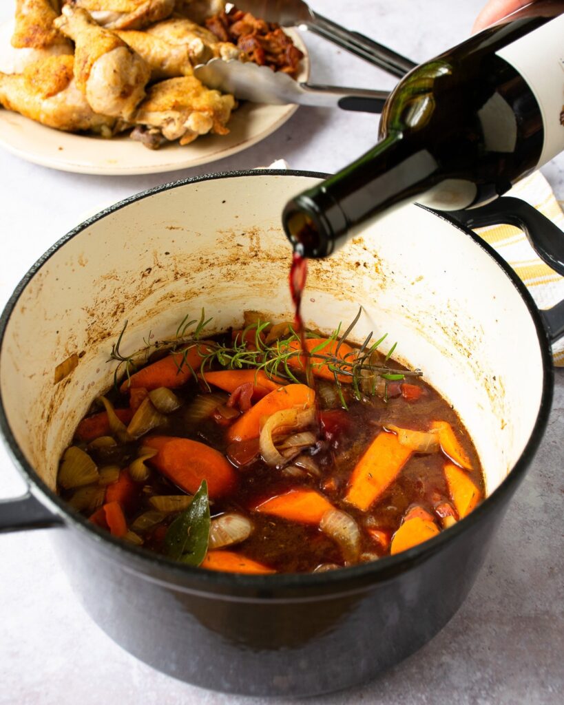Red wine being poured into a Dutch oven with browned chicken on a plate in the background.