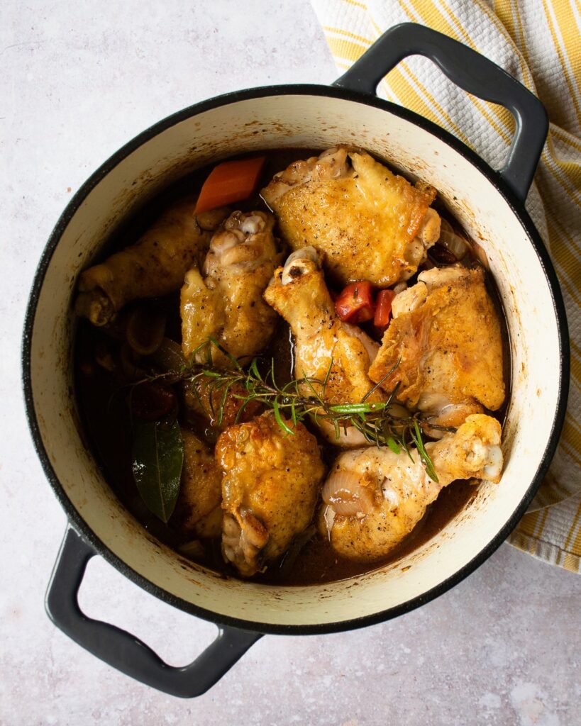 Chicken nestled into braising liquid in a Dutch Oven with a yellow and white striped towel near by.