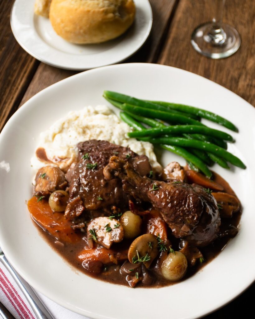 A white plate with Coq au Vin over mashed potatoes with green beans on a wooden table with bread, wine and a napkin.