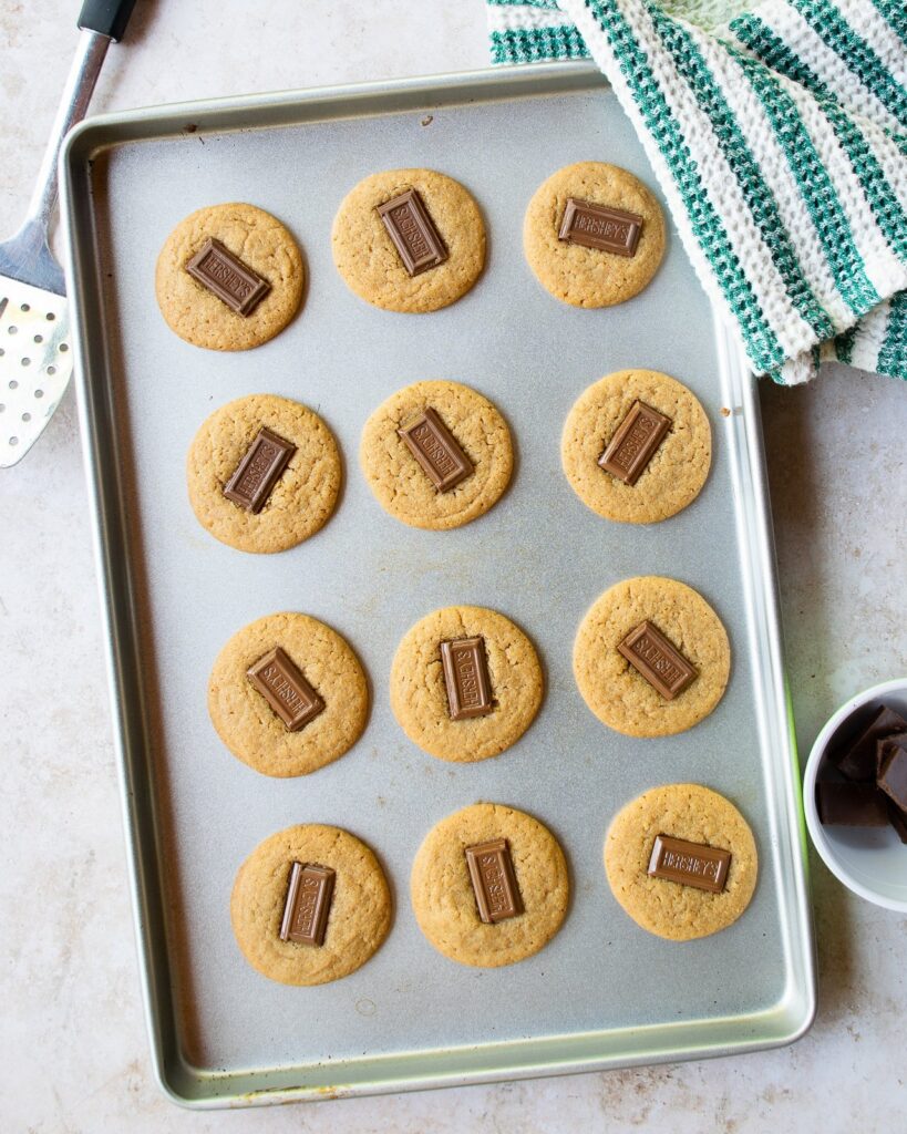 A baking sheet with 12 s'mores cookies, all with a piece of chocolate in the center.
