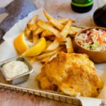 Beer battered fish in a parchment lined basket with air fried french fries, coleslaw, tartar sauce and malt vinegar and a beer in the background.
