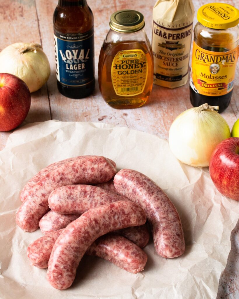 Ingredients on a wooden table - bratwurst, apples, onions, beer, honey, molasses and Worcestershire sauce.