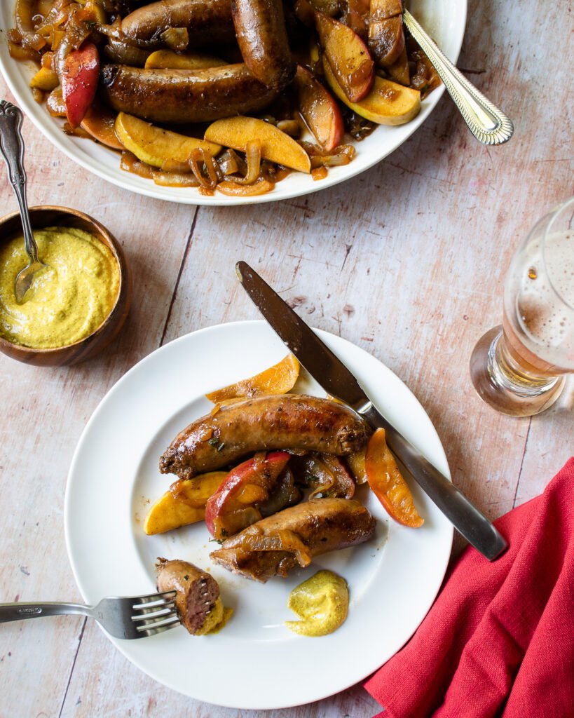 A plate of bratwurst with apples and onions on a table with a family style bowl, mustard and a beer.