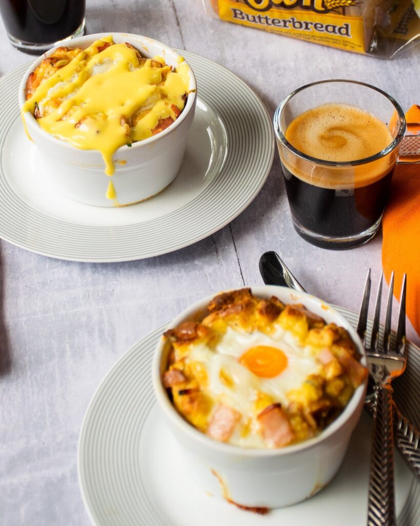 Two ramekins of individual eggs benedict bread puddings - one with Hollandaise sauce and one without - on a table with coffee.
