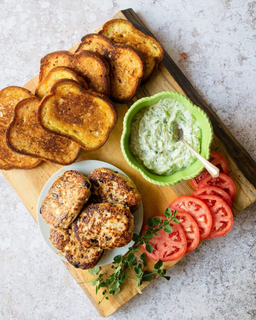 Greek turkey burgers ready to assemble - tomatoes, tzatziki sauce, burger patties, fried bread.