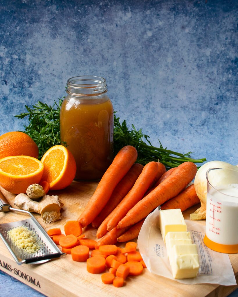 Ingredients on a cutting board with a blue background - carrots, oranges, ginger, chicken stock, cream, butter and an onion.