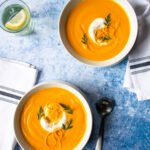 Two bowls of carrot ginger orange soup on a blue table with white napkins and spoons nearby.