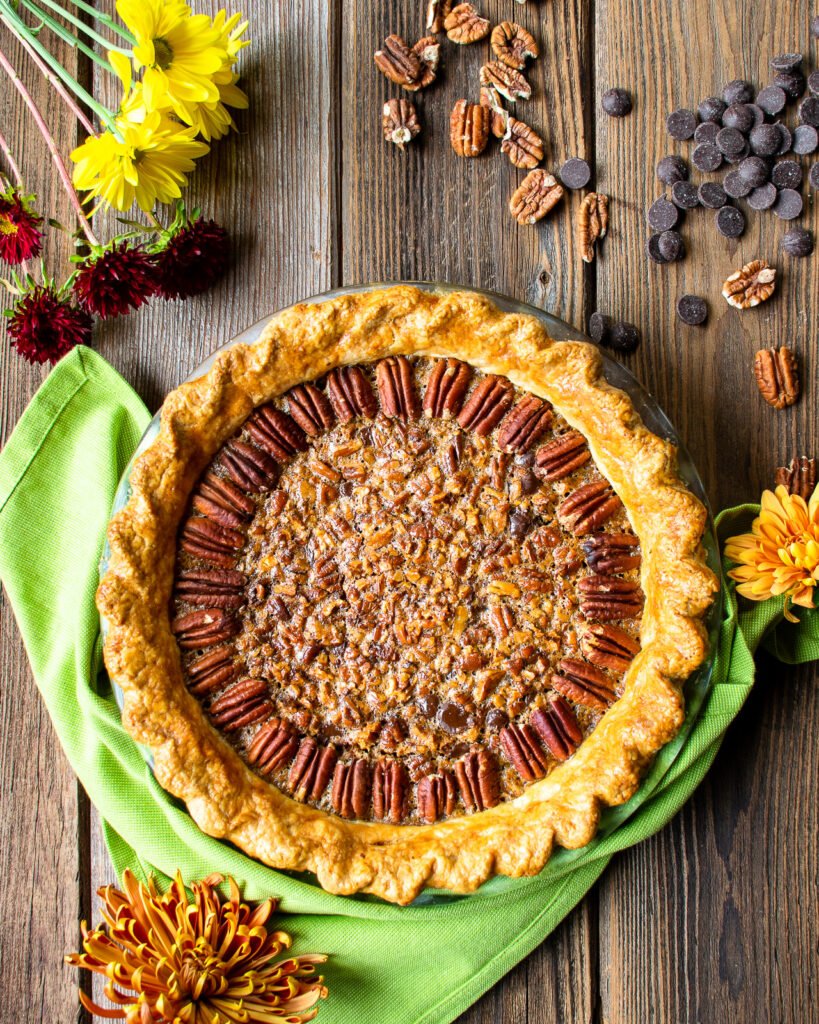 Chocolate Bourbon Pecan Pie, baked and on a wooden table with a green napkin and flowers around.