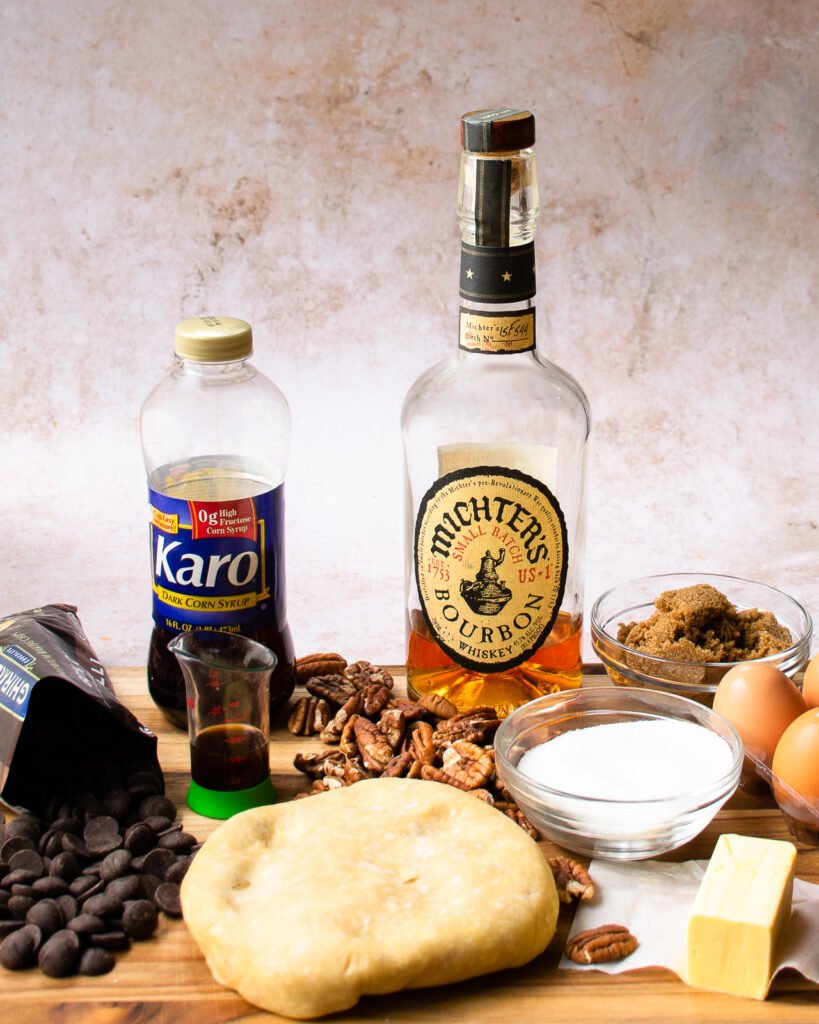 Ingredients on a wooden cutting board - bourbon, pastry, pecans, sugar, butter, eggs, chocolate chips.