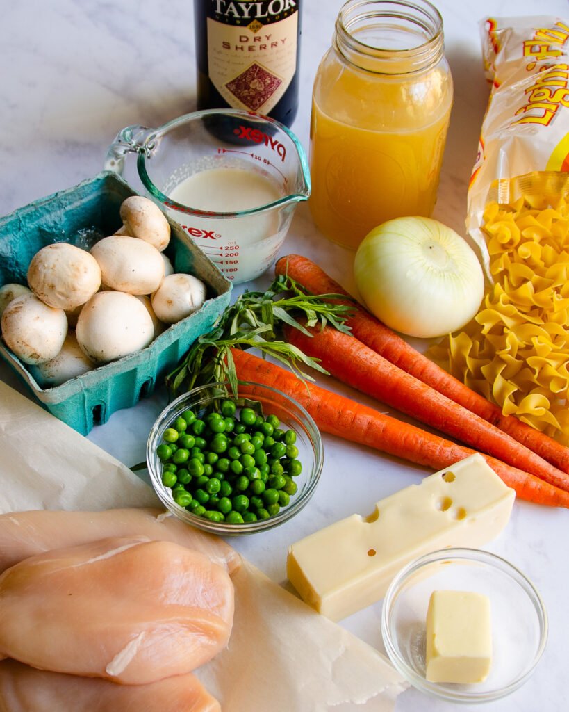 Ingredients on a marble countertop - wine, chicken stock, cream, mushrooms, peas, carrots, onion, pasta, butter, cheese and chicken.