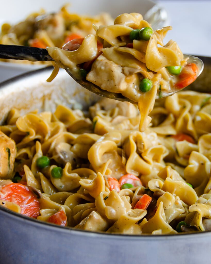 A spoon of creamy tarragon chicken and egg noodles being lifted out of a sauté pan.